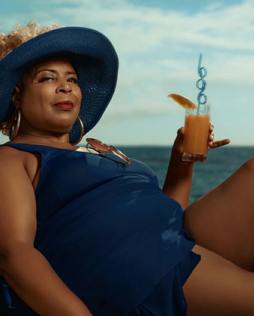 Woman in blue swimsuit and floppy hat relaxing with a cocktail on a sunny day by the ocean, exuding summer vibes and relaxation.